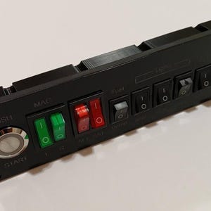 May include: Black control panel with multiple switches and buttons. Features a large silver "START" button and various toggle switches labeled "MAC", "Fuel", and "Lights". Some switches are red and green, others are black. The panel is rectangular.