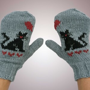 Könnte beinhalten: Ein Paar graue Strickhandschuhe mit schwarzen Katzendesigns und roten Akzenten. Die Handschuhe werden mit ausgestreckten Daumen gezeigt.