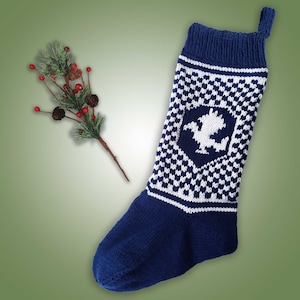May include: A navy blue and white knitted Christmas stocking with a geometric pattern and a white emblem. A sprig of faux pine with red berries and pinecones is to the left of the stocking. The stocking has a loop for hanging.