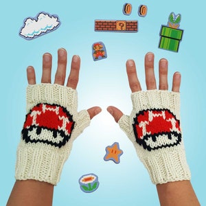 Könnte beinhalten: Ein Paar weiße Fingerlose Handschuhe mit roten und schwarzen Pilzdesigns. Die Handschuhe sind gestrickt und haben ein pixeliges Aussehen. Der Hintergrund zeigt verschiedene pixelartige Super Mario Bros. Charaktere und Objekte.