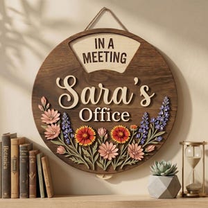 Puede incluir: Un letrero de oficina redondo de madera con el texto "IN A MEETING" en la parte superior y "Sara's Office" en el centro. El letrero está decorado con coloridos detalles florales, que incluyen flores rosas, naranjas y moradas. Se adjunta una cuerda para colgar.