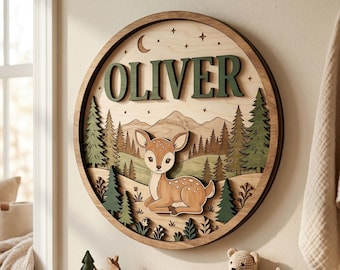May include: A wooden, layered wall art piece with a forest theme. The round sign features the name "OLIVER" in green letters, a deer, trees, and mountains. The sign is approximately 12 inches in diameter and is designed for a nursery or child's room.