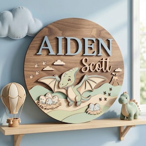 May include: A round wooden sign with the name "AIDEN Scott" in blue and beige lettering. The sign features a 3D design of a cartoon dinosaur, eggs, and a volcano. A plush dinosaur and hot air balloon are on a shelf below.