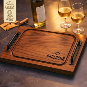 Può includere: Un tagliere rettangolare in legno scuro, con manici neri e un bordo incassato. La tavola è incisa con il nome "ANDERSON" e "CALIFORNIA, USA". L'immagine include anche una bottiglia di vino, bicchieri e cracker.