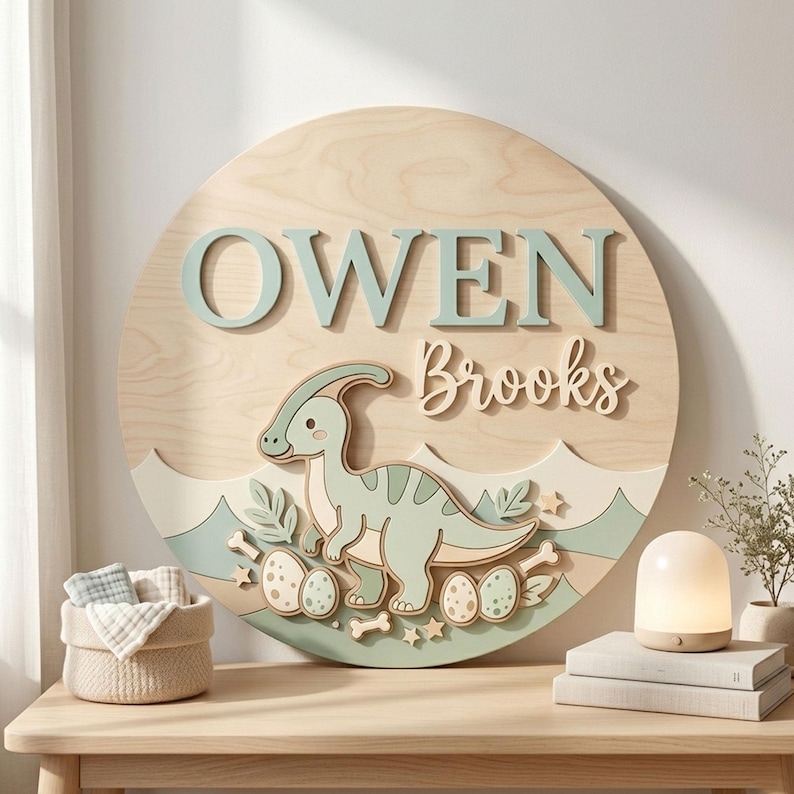 May include: A round wooden sign with the name "OWEN Brooks" in light blue and natural wood tones. A dinosaur illustration is featured below the name, with eggs and stars. The sign is a decorative item.