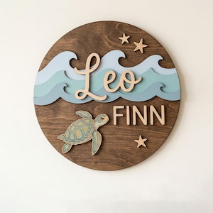 Op de afbeelding: Een ronde, donker gebeitste houten plank met een lichtblauw en blauwgroen golfontwerp, een zeeschildpad en zeesteraccenten. De namen "Leo" en "Finn" staan in een licht houten lettertype. Een nautische wanddecoratie.