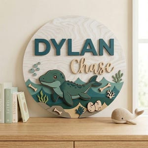 May include: A round wooden sign with the name "DYLAN" in teal letters above "Chase" in a cursive font. The sign features a teal dinosaur illustration, water, bones, and eggs. The background is a light wood grain.