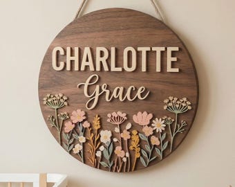 May include: A round wooden sign with the name "CHARLOTTE Grace" in raised letters. The sign features a floral design with pink, yellow, and white flowers. A rope is attached for hanging.