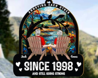 May include: A black and white decorative ornament with the text "ANNOYING EACH OTHER" and "SINCE 1998 AND STILL GOING STRONG." The ornament features a couple sitting in chairs, with a colorful stained-glass background.