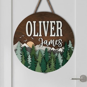 Peut inclure: Panneau rond en bois avec le nom "Oliver James" en lettres blanches. Le panneau présente une scène de forêt en couches avec des arbres verts, des montagnes et un soleil. Il est suspendu par une corde marron.