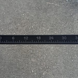 May include: A black ruler with white markings and numbers from 0 to 36 inches. The ruler is rectangular and lies horizontally on a gray surface. The numbers are evenly spaced and clearly visible.