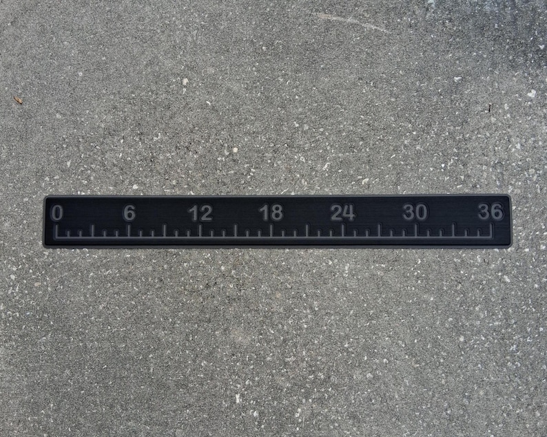 May include: A black ruler with white markings and numbers from 0 to 36 inches. The ruler is rectangular and lies horizontally on a gray surface. The numbers are evenly spaced and clearly visible.