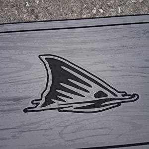 Puede incluir: Un objeto rectangular gris con un dise&ntilde;o de aleta de tibur&oacute;n negra. La aleta est&aacute; delineada en negro y tiene una apariencia texturizada. El fondo tiene un patr&oacute;n de vetas de madera.