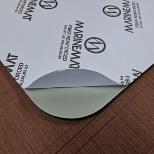Puede incluir: Una l&aacute;mina parcialmente despegada de material Marinemat reforzado con fibra. La capa superior es blanca con texto negro y un logotipo. La capa inferior expuesta es de color verde claro. El material est&aacute; sobre una superficie marr&oacute;n texturizada.