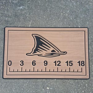 Könnte beinhalten: Fischmessbrett aus Holz mit schwarzem Rand und einem Haifischflossen-Design. Das Brett hat ein Lineal mit Zentimetermarkierungen von 0 bis 45.