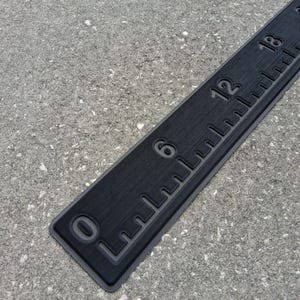 May include: A black ruler with white markings and numbers, including 0, 6, 12, and 18 inches. The ruler has a textured surface and is set against a gray background. The ruler is a measuring tool.