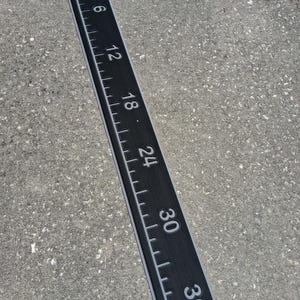 Pode incluir: Uma r&eacute;gua preta com marca&ccedil;&otilde;es e n&uacute;meros brancos, medindo at&eacute; 91,4 cm. A r&eacute;gua tem uma borda cinza e est&aacute; sobre uma superf&iacute;cie cinza. Os n&uacute;meros s&atilde;o claramente vis&iacute;veis, indicando incrementos em cm.