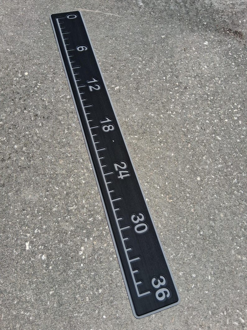 Pode incluir: Uma r&eacute;gua preta com marca&ccedil;&otilde;es e n&uacute;meros brancos, medindo at&eacute; 91,4 cm. A r&eacute;gua tem uma borda cinza e est&aacute; sobre uma superf&iacute;cie cinza. Os n&uacute;meros s&atilde;o claramente vis&iacute;veis, indicando incrementos em cm.