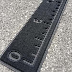 May include: A black ruler with white markings and numbers, including 0, 6, and 12 inches. The ruler has a textured surface and a gray border. The ruler is designed for measuring lengths.