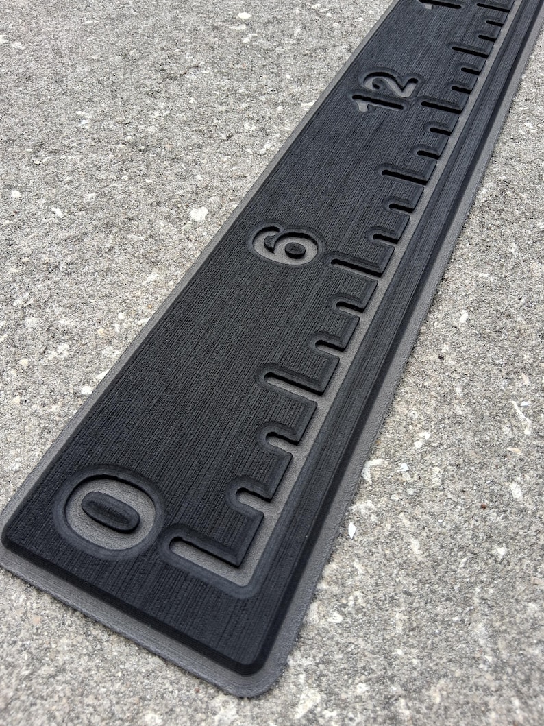 May include: A black ruler with white markings and numbers, including 0, 6, and 12 inches. The ruler has a textured surface and a gray border. The ruler is designed for measuring lengths.