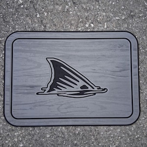 Puede incluir: Alfombra rectangular gris con borde negro y un gr&aacute;fico negro de aleta de tibur&oacute;n emergiendo del agua. La alfombra tiene una textura de vetas de madera.