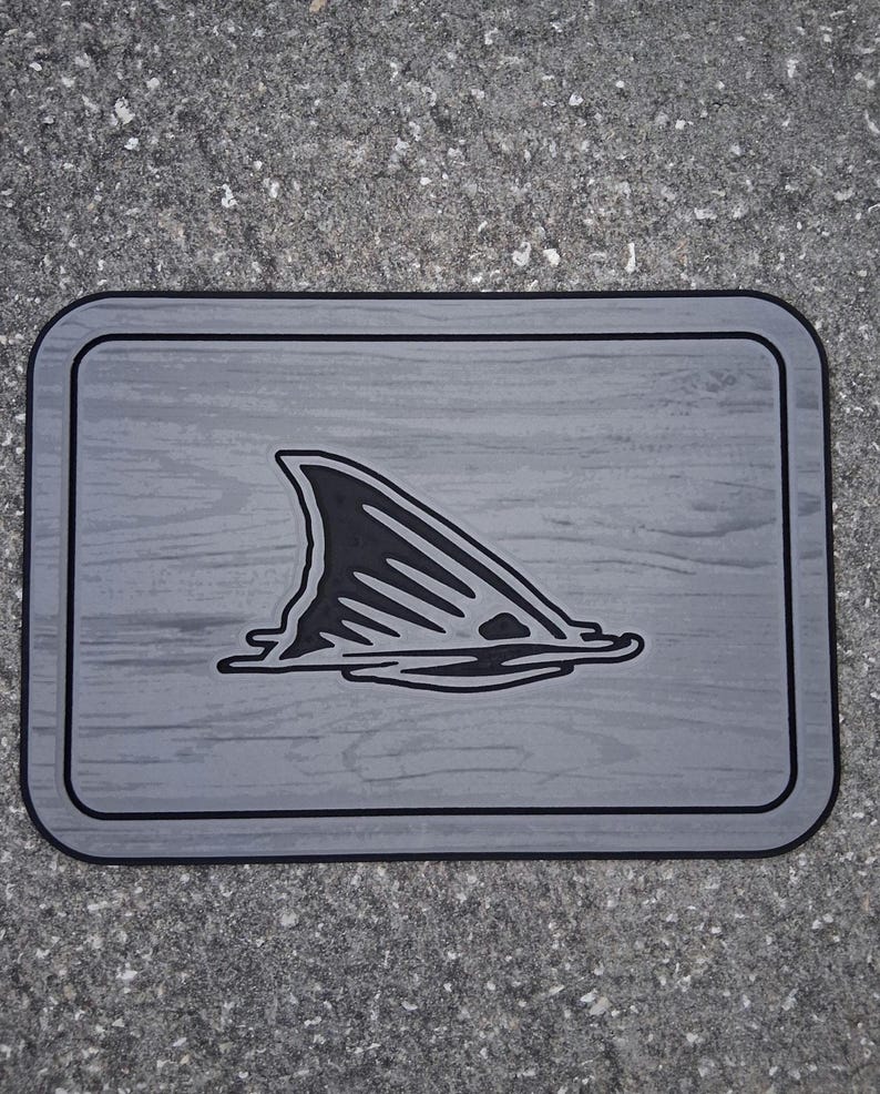 Puede incluir: Alfombra rectangular gris con borde negro y un gr&aacute;fico negro de aleta de tibur&oacute;n emergiendo del agua. La alfombra tiene una textura de vetas de madera.