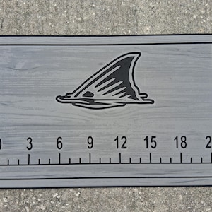 May include: A gray fishing ruler with a black shark fin graphic. The ruler has measurements from 0 to 21 inches, with markings every 3 inches. The background has a wood grain texture.