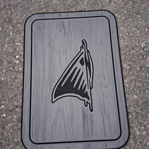 Puede incluir: Una alfombra rectangular gris con un borde negro y una textura de vetas de madera. La alfombra presenta un gr&aacute;fico negro de una forma abstracta estilizada en el centro. La alfombra est&aacute; sobre una superficie gris.