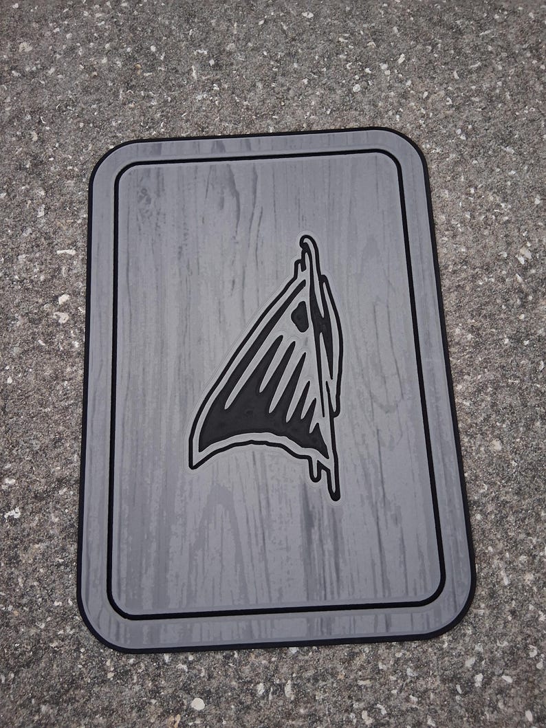 Puede incluir: Una alfombra rectangular gris con un borde negro y una textura de vetas de madera. La alfombra presenta un gr&aacute;fico negro de una forma abstracta estilizada en el centro. La alfombra est&aacute; sobre una superficie gris.