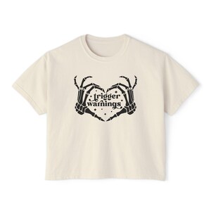 May include: A cream-coloured t-shirt with a black graphic of two skeleton hands forming a heart shape. The words "trigger warnings" are printed in a stylized font within the heart. The shirt has a crew neck and short sleeves.