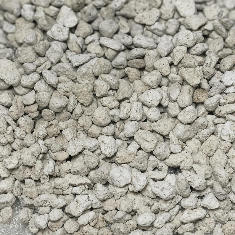 May include: Close-up of a pile of small, irregularly shaped, light gray pumice stones. The stones have a porous texture and vary in size, creating a natural, earthy appearance. The image is well-lit, highlighting the details of each stone.