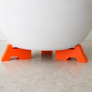 May include: A large, smooth, white, spherical object is elevated by three bright orange, angular, foot-shaped supports. The supports are evenly spaced around the base of the white sphere, providing a stable foundation for the rounded white item.