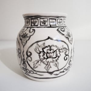 Könnte beinhalten: Eine weiße Keramikvase mit einem schwarzen Blumendesign. Die Vase hat einen runden Körper und einen schmalen Hals. Das Blumendesign zeigt eine große Blume mit Blättern und ein geometrisches Muster um die Oberseite.