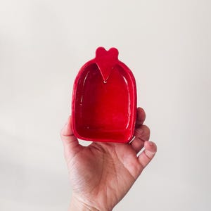 以下が含まれることがあります： 上部にハート型のディテールが付いた、小さく光沢のある赤いセラミック皿。無地の白い背景に対して皿が保持されています。皿は鮮やかで、無地の赤色です。