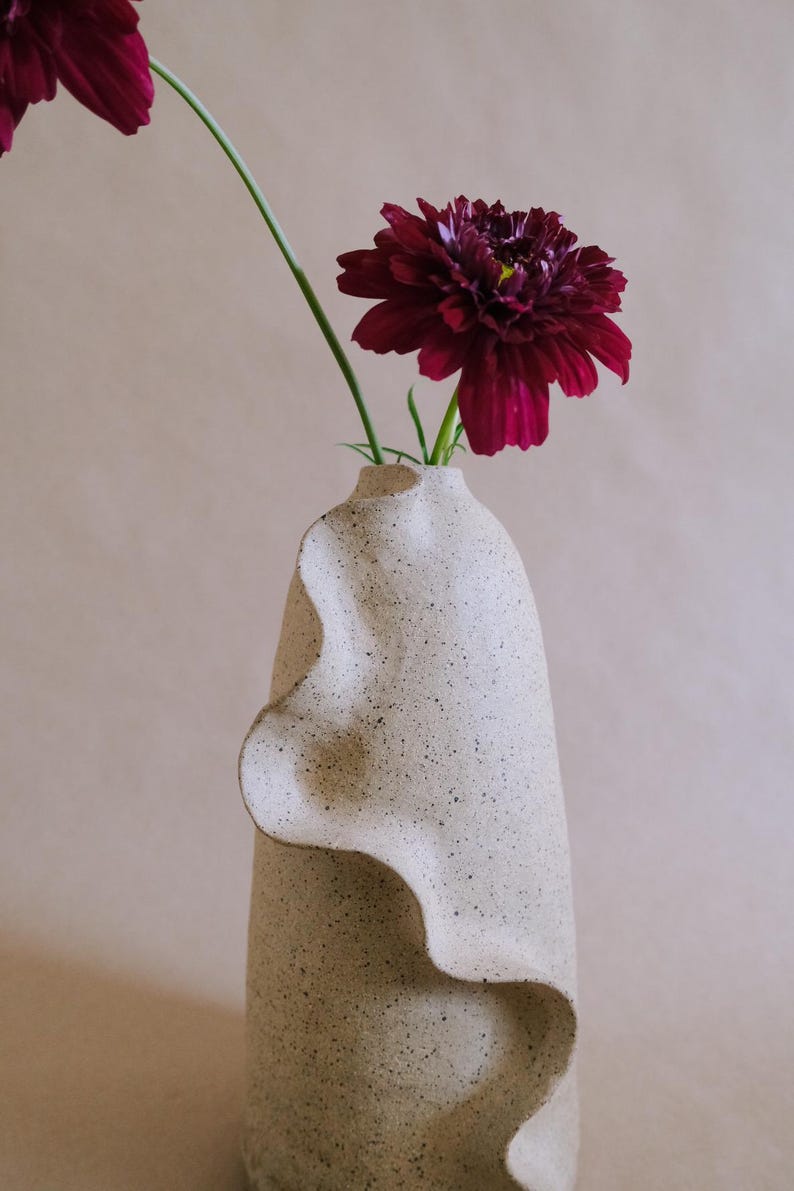 May include: A tall, speckled ceramic vase with a wavy, textured design holds a deep burgundy flower. The flower has a yellow center and green stem. The vase is a neutral color, and the background is a soft beige.