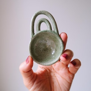 以下が含まれることがあります： 取っ手付きの、小さな斑点のある淡い緑色のセラミック製スプーンレスト。スプーンレストは丸みを帯びたボウルの形をしており、2つのアーチ型のハンドルが付いています。セラミックの作品は、無地の白い背景に対して手で持たれています。