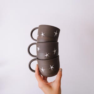 May include: A stack of three dark brown ceramic mugs, each featuring a speckled texture and a pattern of hand-painted white stars. The mugs have rounded bodies and curved handles, arranged vertically to showcase the consistent star design.