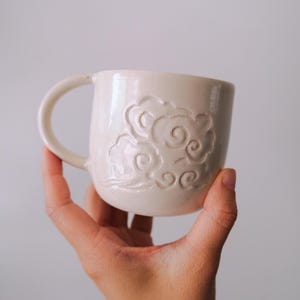 May include: A cream-colored ceramic mug featuring a raised, embossed design of stylized swirling clouds on the exterior surface. The mug has a smooth, glossy finish and a curved handle attached to the side, showcasing a simple, rounded shape.