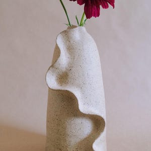 May include: A cream-colored ceramic vase with a wavy, textured design. A deep burgundy flower with green stem is placed inside the vase. The vase has a speckled appearance and a unique, organic shape, suitable for home decor.