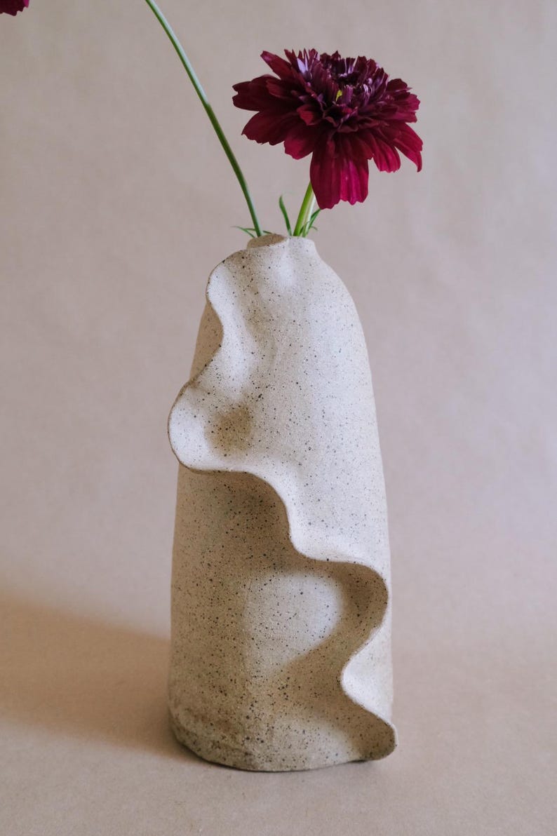 May include: A cream-colored ceramic vase with a wavy, textured design. A deep burgundy flower with green stem is placed inside the vase. The vase has a speckled appearance and a unique, organic shape, suitable for home decor.