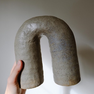 Puede incluir: Escultura de cerámica en forma de U, hecha a mano, con un acabado gris mate moteado. La pieza de arte tiene una forma redondeada con un centro hueco. El color neutro y la forma única la convierten en un acento decorativo.