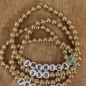 May include: Gold beaded bracelet featuring white letter beads spelling "AMERICA 250 FREEDOM". A small, clear star-shaped charm is attached. The bracelet is displayed on a wooden surface.