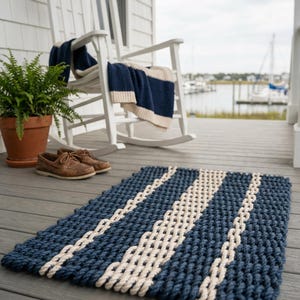 May include: A rectangular blue and white nautical-themed rug with a braided rope design. The rug features alternating stripes of dark blue and off-white. The rug is on a wooden deck, with a white rocking chair and a potted fern in the background.