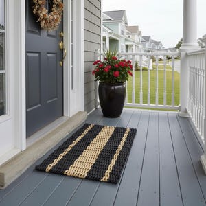 May include: A rectangular doormat featuring a woven pattern of thick black and natural tan rope stripes. The mat is positioned on a gray wooden porch floor in front of a dark gray door with a gold handle and a dried floral wreath hanging above.