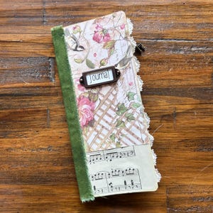 May include: A journal with a floral and lattice patterned cover featuring a green velvet spine, white lace trim, and a metal label holder with the word Journal printed on a white background. The bottom section displays printed musical notation.