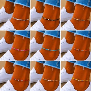 May include: Nine colorful anklets with star and bead accents. Each anklet features a different colored cord, including white, black, pink, turquoise, orange, red, beige, and yellow. The anklets are worn with white sneakers and blue jeans.