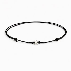 Könnte beinhalten: Ein schwarzes Kordelarmband mit einem kleinen, silberfarbenen Sternanhänger. Das Armband ist an jedem Ende und in der Mitte geknotet, wobei sich der Sternanhänger in der Mitte befindet. Das Armband wird vor einem weißen Hintergrund gezeigt.