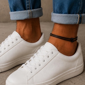 Puede incluir: Primer plano de los tobillos de una persona que lleva una tobillera negra de doble hebra. La persona lleva zapatillas blancas y vaqueros azules con los puños doblados. El fondo es gris neutro.