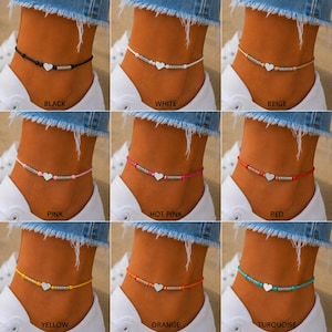 May include: A collection of nine anklets in black, white, beige, pink, hot pink, red, yellow, orange, and turquoise. Each anklet features a central silver-toned heart charm flanked by small metallic beads on a thin, coloured cord string.
