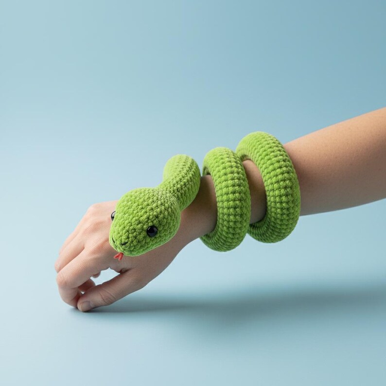 K&ouml;nnte beinhalten: Ein gr&uuml;nes geh&auml;keltes Schlangenarmband mit roter Zunge und schwarzen Augen. Der K&ouml;rper der Schlange wickelt sich um das Handgelenk und den Arm. Der Hintergrund ist einfarbig hellblau.